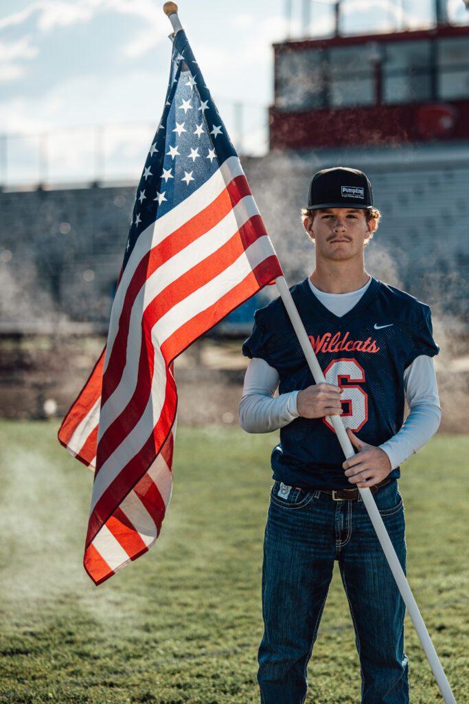filer idaho southern idaho football sportrait Young man in a Wildcats football jersey and cap stands on a field holding a large American flag, with bleachers and sunlight in the background.