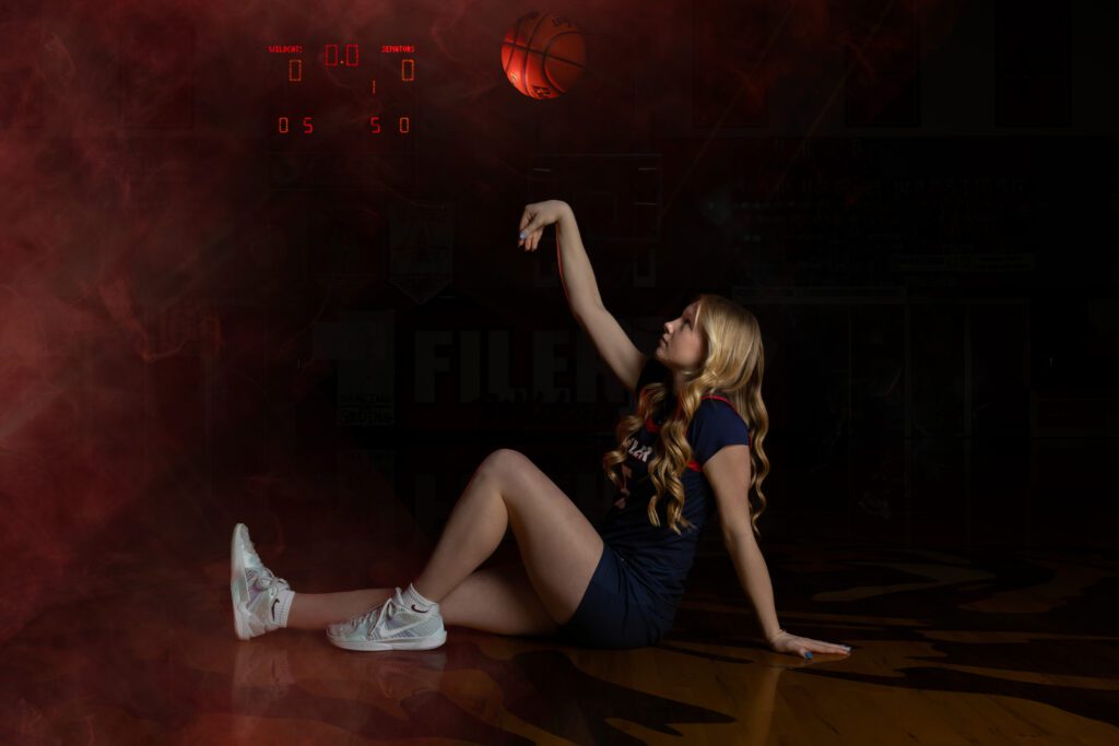 filer idaho southern idaho basketball sportrait A woman in a navy athletic outfit sits on a basketball court, reaching toward a hoop amid red smoke and dramatic, moody lighting.